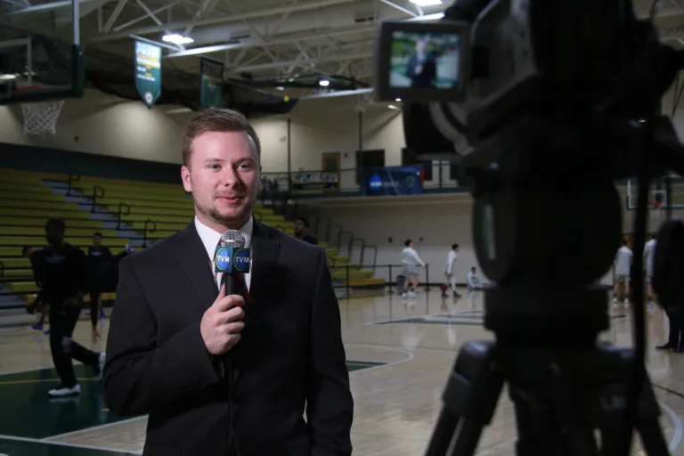 A sports journalist student stands in front of a camera while holding a microphone. He stands in a gymnasium while basketball players are playing behind him.